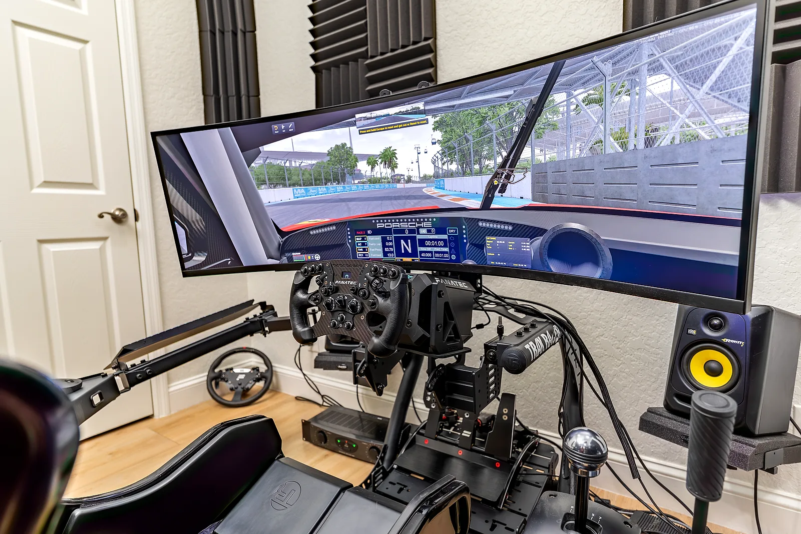 A Fanatec sim-racing cockpit: a Podium DD1 wheelbase and Formula wheel on a Trak Racer TR8 Pro frame with ClubSport V3 pedals, in front of a curved 49-inch ultrawide running iRacing, beside studio monitors and acoustic panels